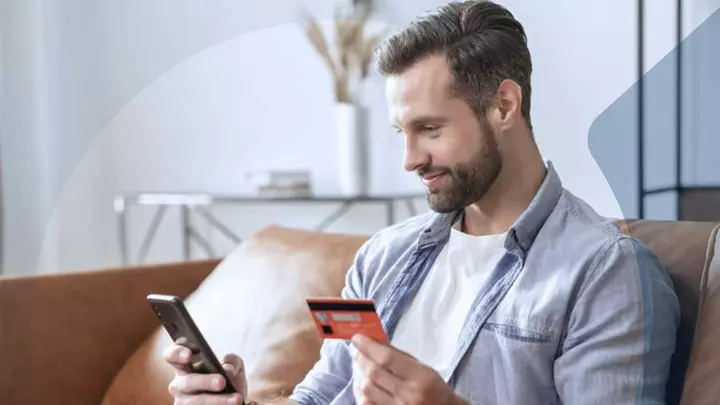 Man sitting on a sofa holding a credit card and smartphone, symbolizing credit card data security under PCI-DSS guidelines.